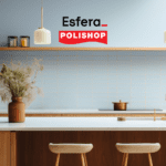 Uma cozinha moderna com uma ilha central em madeira clara, decorada com um vaso de flores secas. No fundo, uma parede com azulejos claros e prateleiras minimalistas. As logos "Esfera" e "Polishop" estão posicionadas na parte superior da imagem.