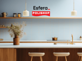 Uma cozinha moderna com uma ilha central em madeira clara, decorada com um vaso de flores secas. No fundo, uma parede com azulejos claros e prateleiras minimalistas. As logos "Esfera" e "Polishop" estão posicionadas na parte superior da imagem.