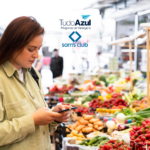 Uma mulher está em uma feira, cercada por bancas de frutas e legumes frescos. Ela olha para seu celular enquanto faz compras. As logos do "TudoAzul Programa de Vantagens" e "Sam's Club" estão posicionadas na parte superior da imagem.