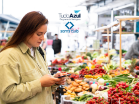 Uma mulher está em uma feira, cercada por bancas de frutas e legumes frescos. Ela olha para seu celular enquanto faz compras. As logos do "TudoAzul Programa de Vantagens" e "Sam's Club" estão posicionadas na parte superior da imagem.