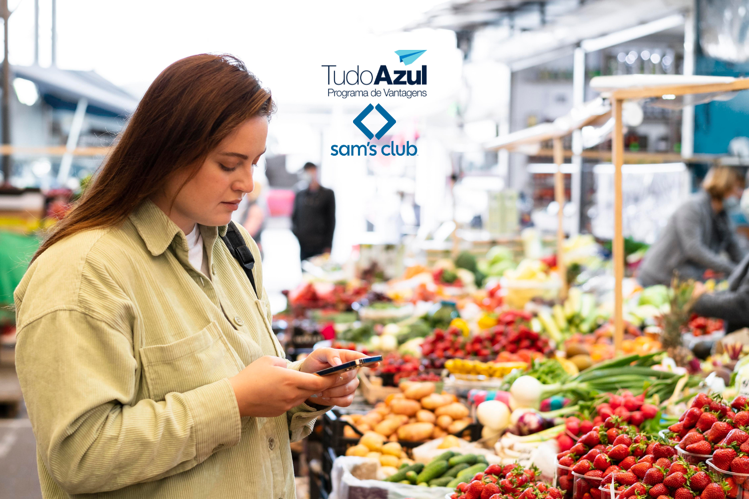Uma mulher está em uma feira, cercada por bancas de frutas e legumes frescos. Ela olha para seu celular enquanto faz compras. As logos do "TudoAzul Programa de Vantagens" e "Sam's Club" estão posicionadas na parte superior da imagem.