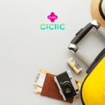 Uma mala amarela com um chapéu de palha e uma miniatura de avião estão ao lado de um passaporte, uma câmera vintage, passagens aéreas e uma pequena quantia de dinheiro em dólar enrolada. As logos da "Livelo" e "Ciclic" estão posicionadas acima da imagem.