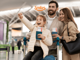Uma família feliz no aeroporto. Uma menina empolgada aponta para algo à frente enquanto é empurrada em um carrinho de bagagem por seus pais, que sorriem ao lado dela. Todos estão segurando passaportes e passagens aéreas. A logo da "Smiles" está posicionada acima da imagem.