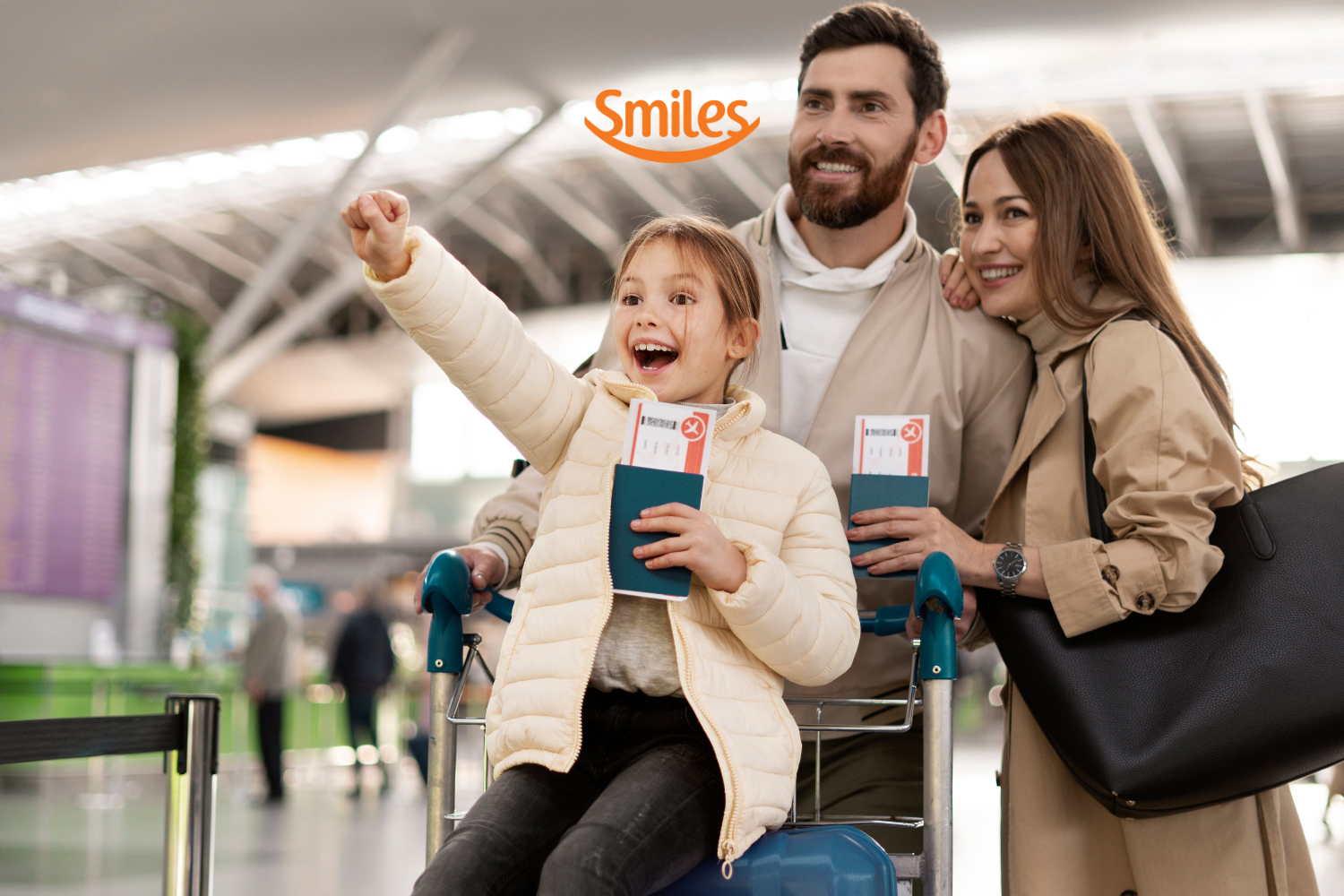 Uma família feliz no aeroporto. Uma menina empolgada aponta para algo à frente enquanto é empurrada em um carrinho de bagagem por seus pais, que sorriem ao lado dela. Todos estão segurando passaportes e passagens aéreas. A logo da "Smiles" está posicionada acima da imagem.