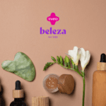 Uma composição de produtos de beleza e cuidados pessoais está disposta sobre uma superfície bege. Entre os itens, há frascos de vidro e madeira, um rolo de jade, uma ferramenta de massagem facial em formato de coração e uma flor branca. No centro da imagem, acima, estão as logos da Livelo e Beleza na Web.