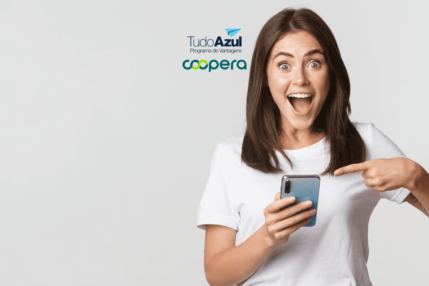 Uma mulher com expressão de surpresa e empolgação segura um smartphone na mão direita enquanto aponta para ele com a mão esquerda. Ela veste uma camiseta branca e tem cabelos castanhos. Acima dela, estão as logos do TudoAzul Programa de Vantagens e da Coopera.