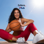 Uma mulher sorridente, usando roupa esportiva, está sentada ao ar livre segurando uma bola de basquete. Ela veste uma calça legging vermelha, top branco e tênis branco, com o mar ao fundo sob um céu claro. Acima dela, estão as logos da Esfera e Fila.