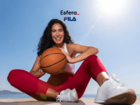 Uma mulher sorridente, usando roupa esportiva, está sentada ao ar livre segurando uma bola de basquete. Ela veste uma calça legging vermelha, top branco e tênis branco, com o mar ao fundo sob um céu claro. Acima dela, estão as logos da Esfera e Fila.