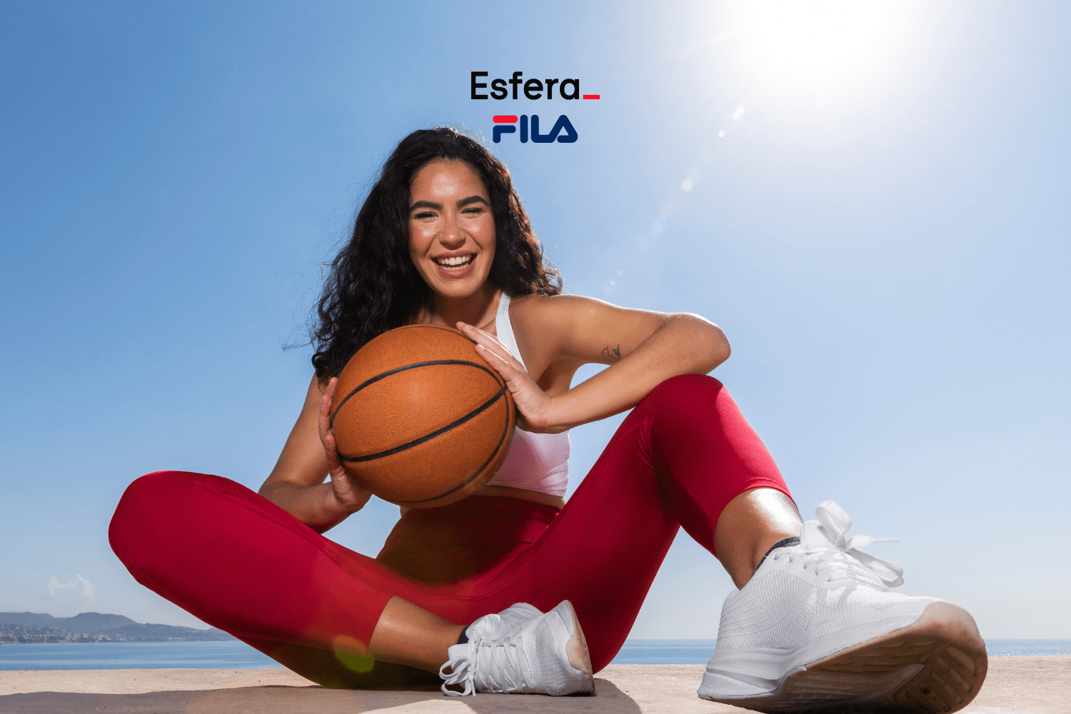 Uma mulher sorridente, usando roupa esportiva, está sentada ao ar livre segurando uma bola de basquete. Ela veste uma calça legging vermelha, top branco e tênis branco, com o mar ao fundo sob um céu claro. Acima dela, estão as logos da Esfera e Fila.