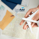 Uma mão segura um modelo de avião branco sobre uma mesa, onde também há um mapa e uma fotografia de praia com a palavra "PARADISE" escrita na areia. Acima, está a logo do programa de vantagens TudoAzul
