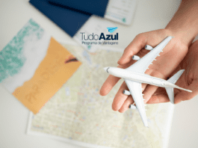 Uma mão segura um modelo de avião branco sobre uma mesa, onde também há um mapa e uma fotografia de praia com a palavra "PARADISE" escrita na areia. Acima, está a logo do programa de vantagens TudoAzul