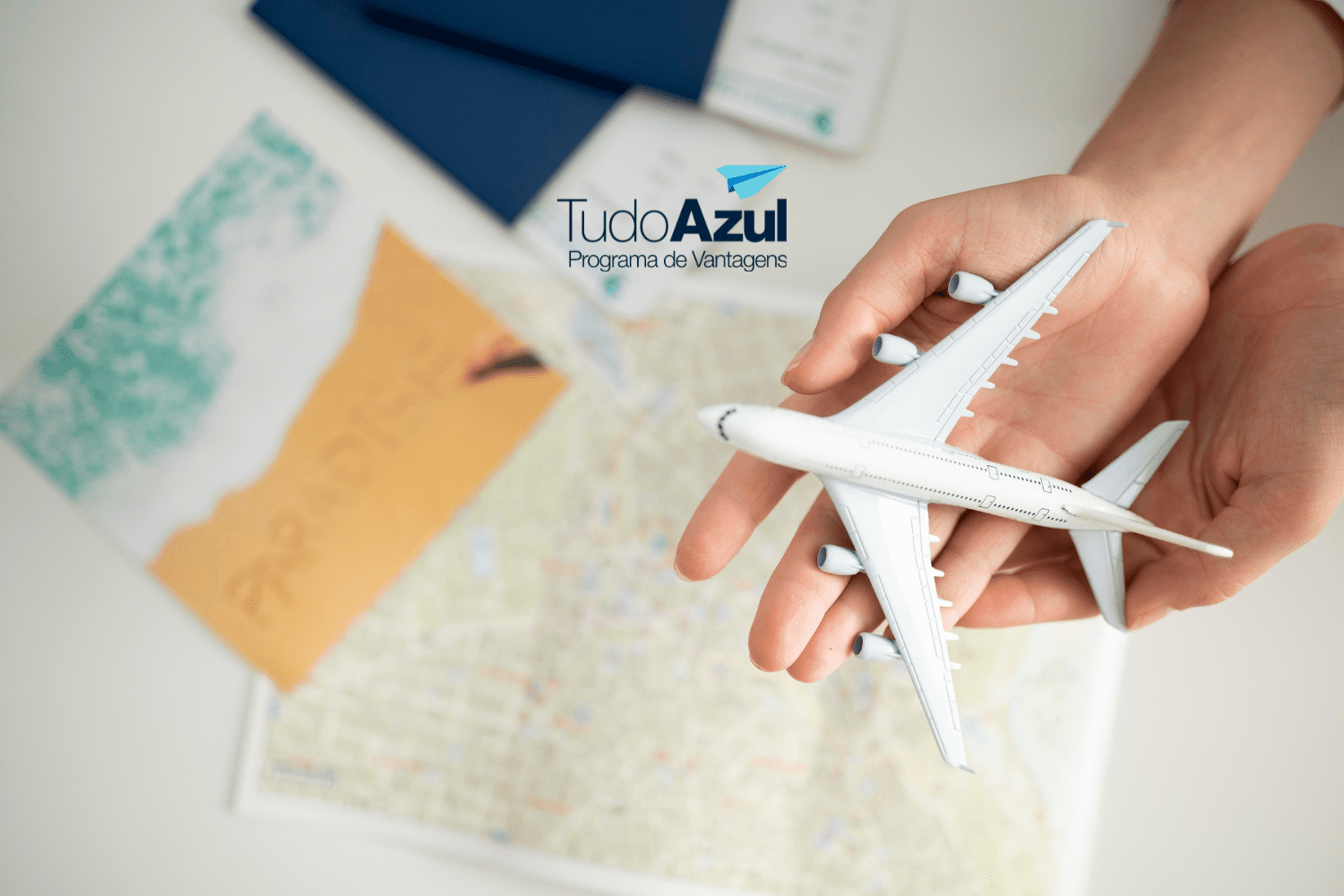 Uma mão segura um modelo de avião branco sobre uma mesa, onde também há um mapa e uma fotografia de praia com a palavra "PARADISE" escrita na areia. Acima, está a logo do programa de vantagens TudoAzul
