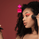Uma mulher com cabelo cacheado aplica maquiagem com um pincel no rosto enquanto se olha em um espelho de mão. Ela está com sombras em tons quentes nos olhos. Acima, estão as logos da Livelo e da Avon.