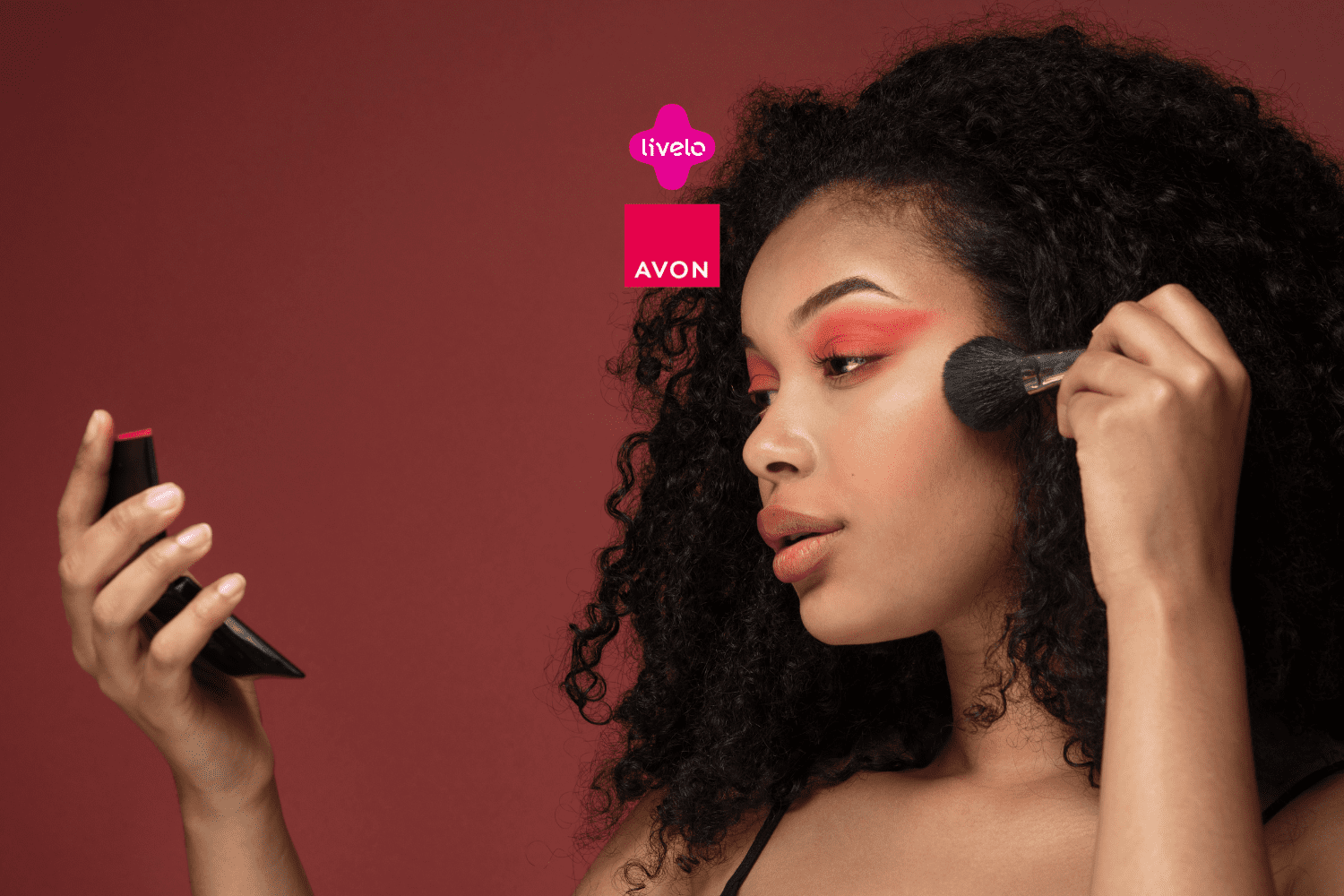 Uma mulher com cabelo cacheado aplica maquiagem com um pincel no rosto enquanto se olha em um espelho de mão. Ela está com sombras em tons quentes nos olhos. Acima, estão as logos da Livelo e da Avon.
