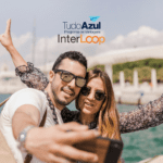 **Texto alternativo:** Um casal sorridente tira uma selfie em um local ensolarado, próximo a uma área com água e um fundo urbano. Ambos estão usando óculos de sol e parecem estar se divertindo. Acima da imagem, estão as logos do programa de vantagens TudoAzul e da empresa InterLoop.