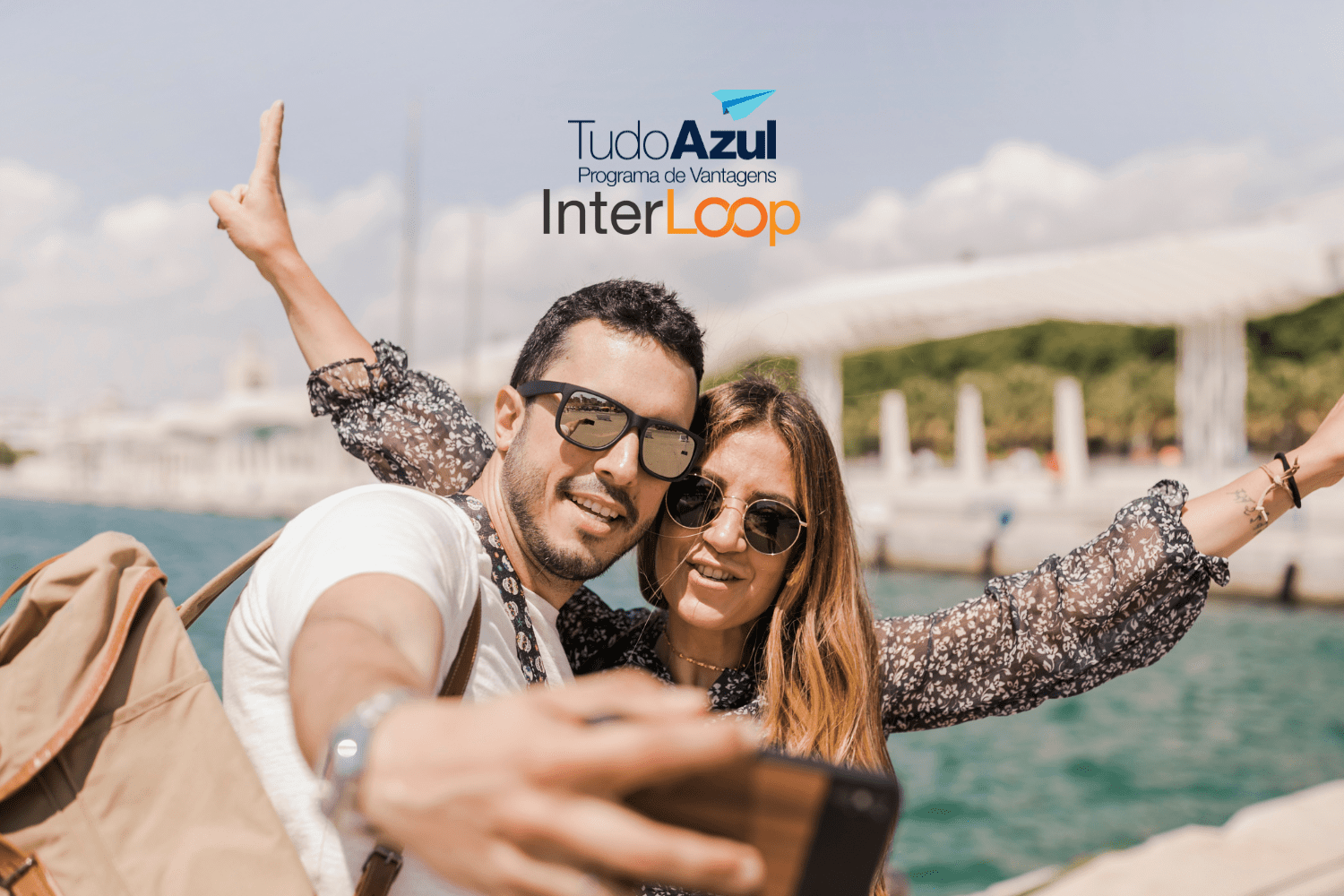 **Texto alternativo:** Um casal sorridente tira uma selfie em um local ensolarado, próximo a uma área com água e um fundo urbano. Ambos estão usando óculos de sol e parecem estar se divertindo. Acima da imagem, estão as logos do programa de vantagens TudoAzul e da empresa InterLoop.