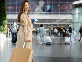 Uma mulher sorridente, vestida com um casaco bege, puxa uma mala de rodinhas em um saguão de aeroporto movimentado. Ela segura passagens aéreas e olha para trás enquanto caminha. Acima da imagem, está a logo do programa de vantagens TudoAzul.