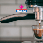 Imagem de uma máquina de café expresso em close-up, enquanto extrai um café em um pequeno copo de vidro. Acima da imagem, estão as logos da Livelo e das Casas Bahia.