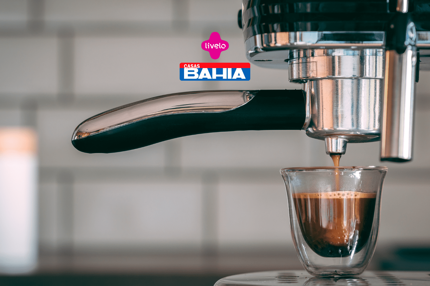 Imagem de uma máquina de café expresso em close-up, enquanto extrai um café em um pequeno copo de vidro. Acima da imagem, estão as logos da Livelo e das Casas Bahia.