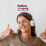 Imagem de uma mulher jovem sorrindo, usando fones de ouvido brancos e fazendo um sinal de "joinha" com as duas mãos. Acima da imagem, estão as logos da Esfera e das Casas Bahia.