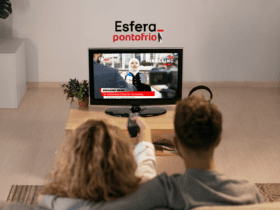 **Texto alternativo:** Imagem de um casal assistindo televisão em uma sala de estar. A TV está transmitindo uma reportagem ao vivo com o título "Breaking News". Acima da TV, estão as logos da Esfera e do Ponto Frio.