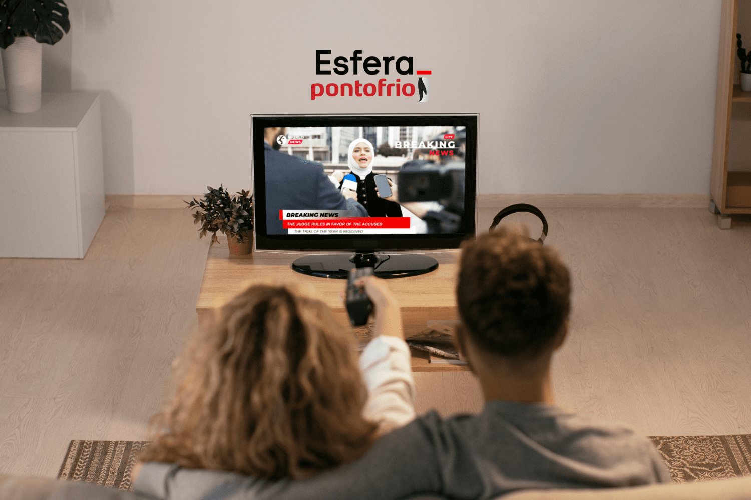**Texto alternativo:** Imagem de um casal assistindo televisão em uma sala de estar. A TV está transmitindo uma reportagem ao vivo com o título "Breaking News". Acima da TV, estão as logos da Esfera e do Ponto Frio.