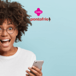 **Texto alternativo:** Imagem de uma mulher jovem e animada, com cabelos cacheados e volumosos, usando óculos redondos e segurando um smartphone. Ela está sorrindo amplamente com expressão de surpresa. Ao fundo, um fundo azul claro e, acima da mulher, estão as logos da Livelo e Ponto Frio.