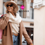 **Texto alternativo:** Imagem de uma mulher sorridente usando um chapéu e óculos escuros, vestindo um casaco marrom e segurando várias sacolas de compras coloridas. Ao fundo, há uma fachada de um prédio elegante. Acima da mulher, estão as logos da Livelo e Renner.