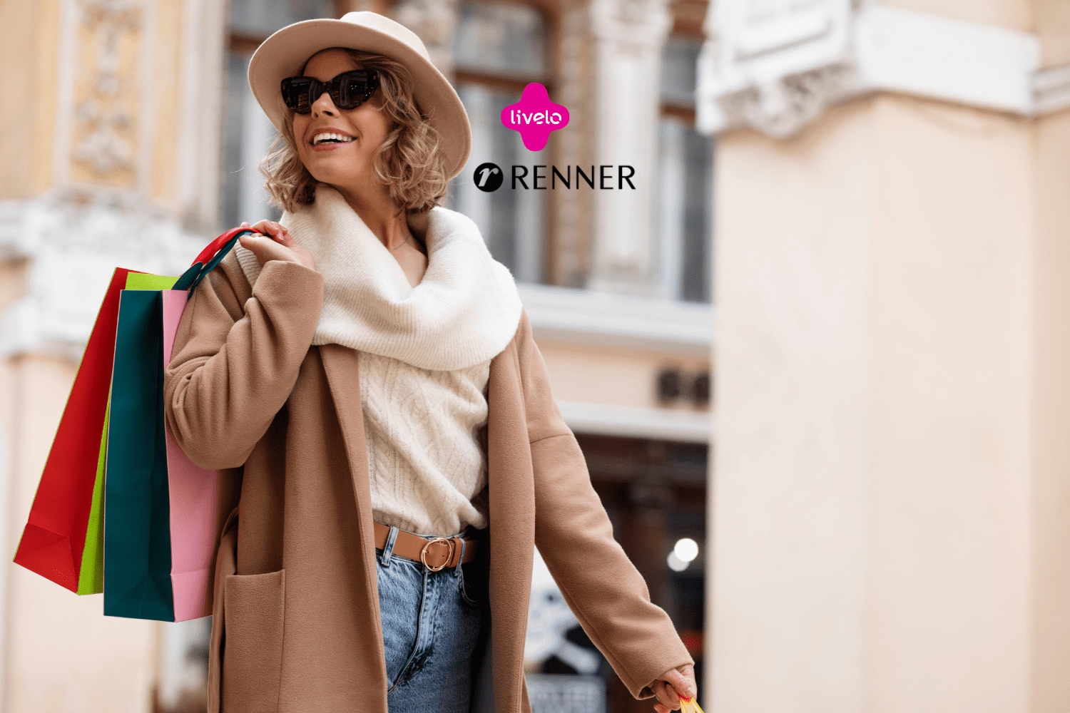 **Texto alternativo:** Imagem de uma mulher sorridente usando um chapéu e óculos escuros, vestindo um casaco marrom e segurando várias sacolas de compras coloridas. Ao fundo, há uma fachada de um prédio elegante. Acima da mulher, estão as logos da Livelo e Renner.