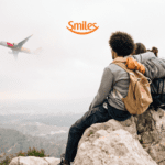 Grupo de amigos sentados em uma rocha, apreciando a vista de um avião que passa ao fundo, envolto por nuvens. Logotipo da Smiles ao topo da imagem.