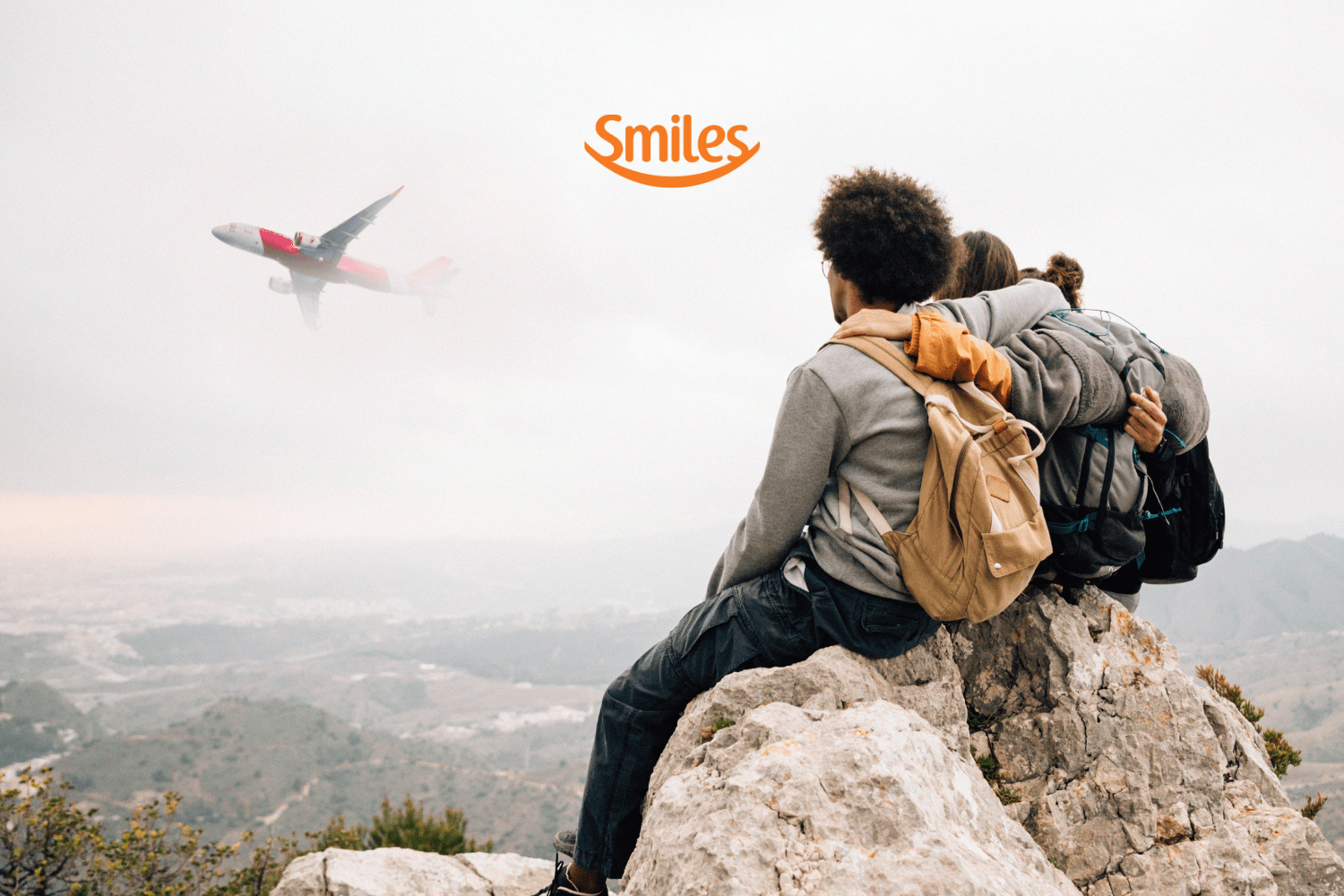 Grupo de amigos sentados em uma rocha, apreciando a vista de um avião que passa ao fundo, envolto por nuvens. Logotipo da Smiles ao topo da imagem.