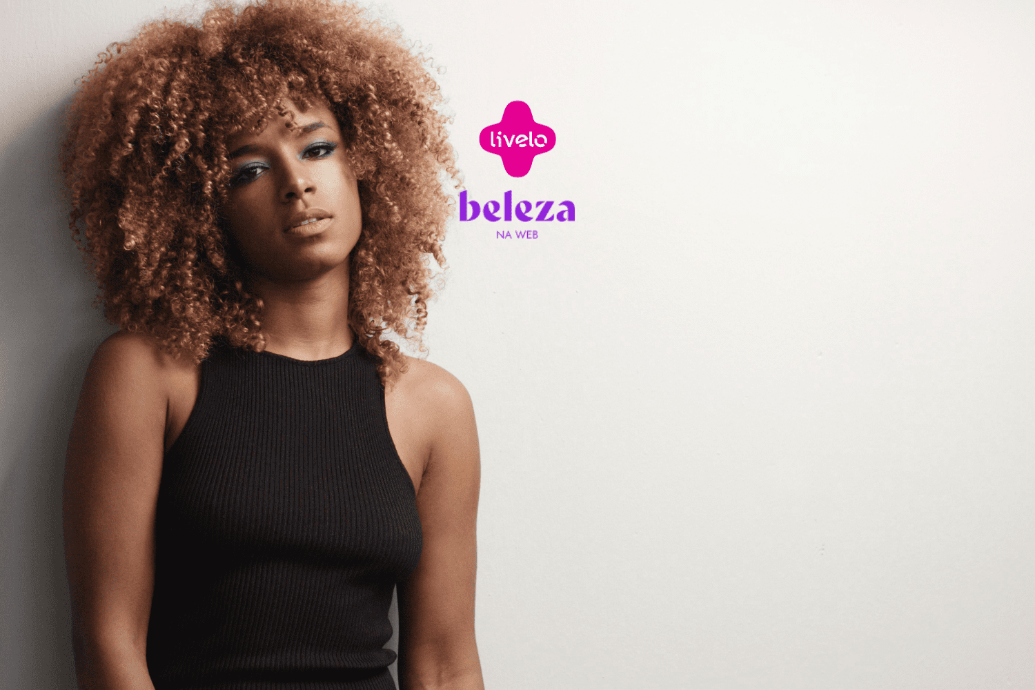Descrição alternativa curta: Mulher com cabelo cacheado volumoso e maquiagem destacada, olhando para a câmera. Logotipo da Livelo e Beleza na Web acima.