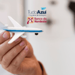 Mão segurando um avião em miniatura com os logotipos do programa de vantagens TudoAzul e do Banco do Nordeste ao fundo.