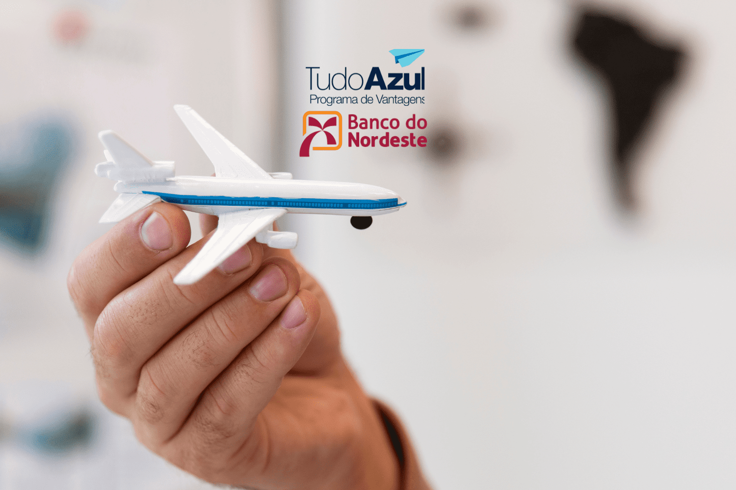 Mão segurando um avião em miniatura com os logotipos do programa de vantagens TudoAzul e do Banco do Nordeste ao fundo.