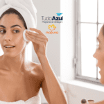 Mulher em um ambiente de banheiro aplicando um produto na pele com algodão, enquanto usa toalha na cabeça. Ao lado, os logotipos do programa de vantagens TudoAzul e da marca Natura.