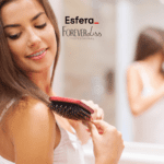 Descrição alternativa curta: Mulher sorridente penteando o cabelo com uma escova vermelha, em frente a um espelho. Na parte superior da imagem, os logotipos da Esfera e Forever Liss Professional.