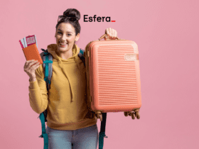 Descrição alternativa curta: Mulher sorridente, vestindo moletom amarelo e mochila verde, segurando uma mala cor de pêssego e um passaporte com passagens. Ao fundo, um fundo rosa e o logotipo da Esfera na parte superior da imagem.