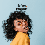 Uma jovem mulher de óculos de sol com armação transparente e lentes laranja, com cabelo cacheado volumoso, usando um suéter amarelo claro, posando contra um fundo azul claro. Acima dela, está o logotipo da Esfera e Insider.