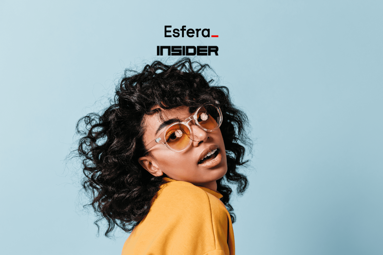 Uma jovem mulher de óculos de sol com armação transparente e lentes laranja, com cabelo cacheado volumoso, usando um suéter amarelo claro, posando contra um fundo azul claro. Acima dela, está o logotipo da Esfera e Insider.
