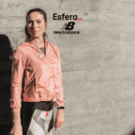 Na imagem, uma mulher está em pé ao lado de uma parede de concreto, vestindo uma jaqueta rosa e roupas de treino, com fones de ouvido. No canto superior da imagem, estão as logomarcas "Esfera_" em preto com um sublinhado vermelho e "New Balance" em preto, representando uma parceria entre as marcas.
