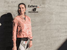 Na imagem, uma mulher está em pé ao lado de uma parede de concreto, vestindo uma jaqueta rosa e roupas de treino, com fones de ouvido. No canto superior da imagem, estão as logomarcas "Esfera_" em preto com um sublinhado vermelho e "New Balance" em preto, representando uma parceria entre as marcas.