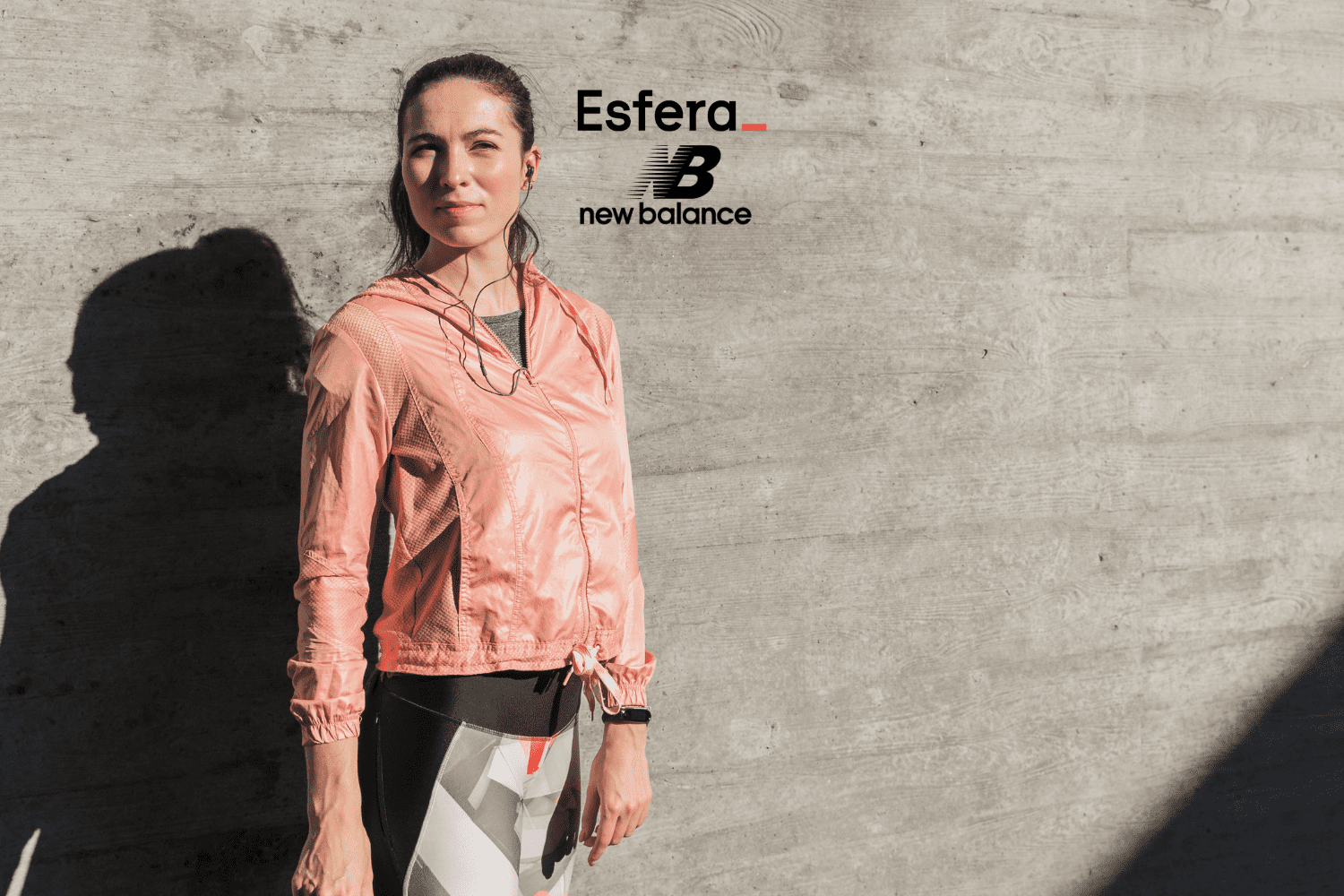 Na imagem, uma mulher está em pé ao lado de uma parede de concreto, vestindo uma jaqueta rosa e roupas de treino, com fones de ouvido. No canto superior da imagem, estão as logomarcas "Esfera_" em preto com um sublinhado vermelho e "New Balance" em preto, representando uma parceria entre as marcas.