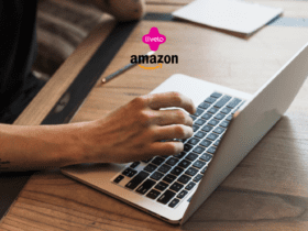 Na imagem, uma pessoa está usando um laptop sobre uma mesa de madeira, digitando no teclado. No canto superior central da imagem, estão as logomarcas "Livelo" em rosa com o texto em branco e "Amazon" em preto com a seta amarela característica abaixo, indicando uma colaboração ou promoção entre essas duas marcas.