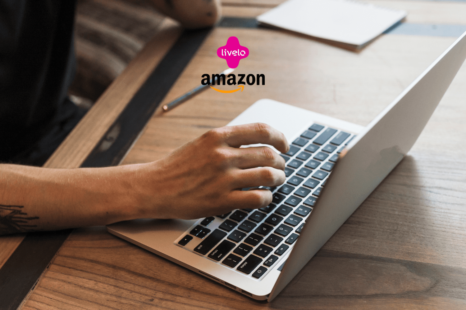 Na imagem, uma pessoa está usando um laptop sobre uma mesa de madeira, digitando no teclado. No canto superior central da imagem, estão as logomarcas "Livelo" em rosa com o texto em branco e "Amazon" em preto com a seta amarela característica abaixo, indicando uma colaboração ou promoção entre essas duas marcas.