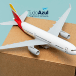 Na imagem, há um modelo de avião em miniatura pousado sobre uma caixa de papelão. O fundo é de cor azul clara, o que destaca o avião e a caixa. No canto superior direito da imagem, está o logotipo do programa de vantagens "TudoAzul", com o texto em azul escuro e um ícone de um avião de papel azul acima do texto. A imagem sugere uma associação entre o programa de fidelidade TudoAzul e a ideia de viagem ou transporte.