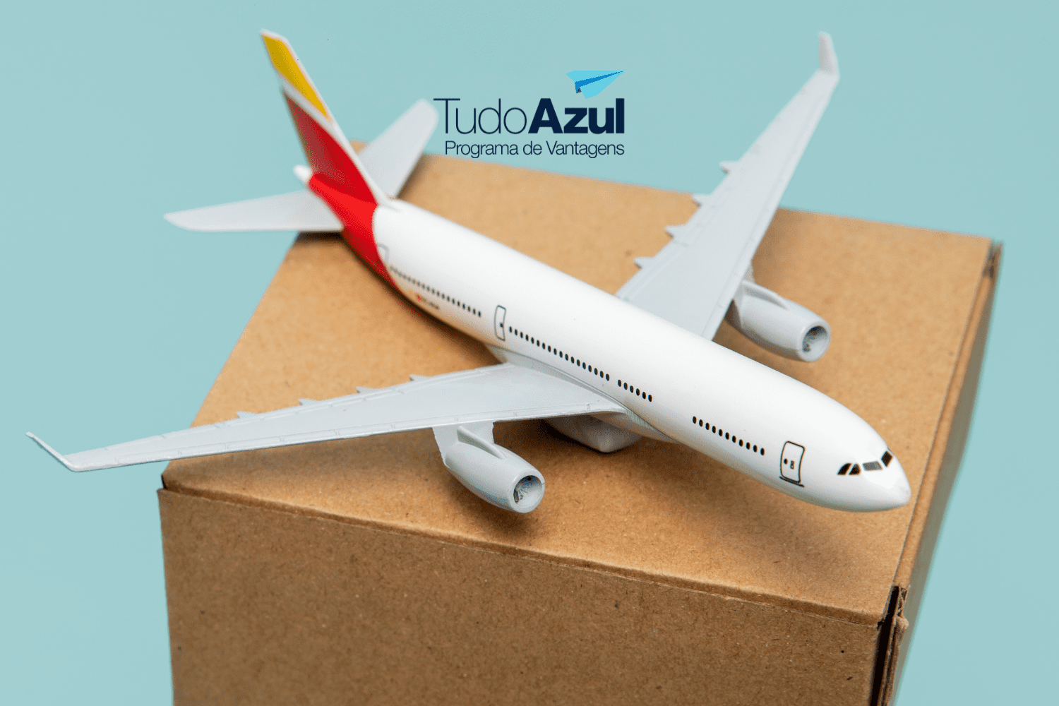 Na imagem, há um modelo de avião em miniatura pousado sobre uma caixa de papelão. O fundo é de cor azul clara, o que destaca o avião e a caixa. No canto superior direito da imagem, está o logotipo do programa de vantagens "TudoAzul", com o texto em azul escuro e um ícone de um avião de papel azul acima do texto. A imagem sugere uma associação entre o programa de fidelidade TudoAzul e a ideia de viagem ou transporte.