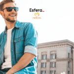 Homem branco sorrindo, usando óculos de sol, jaqueta jeans e camiseta branca, sentado ao ar livre em frente a um prédio histórico. Acima dele estão os logos da Esfera e da Natura, indicando uma colaboração entre as marcas.