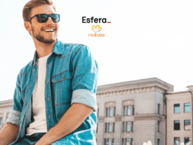 Homem branco sorrindo, usando óculos de sol, jaqueta jeans e camiseta branca, sentado ao ar livre em frente a um prédio histórico. Acima dele estão os logos da Esfera e da Natura, indicando uma colaboração entre as marcas.