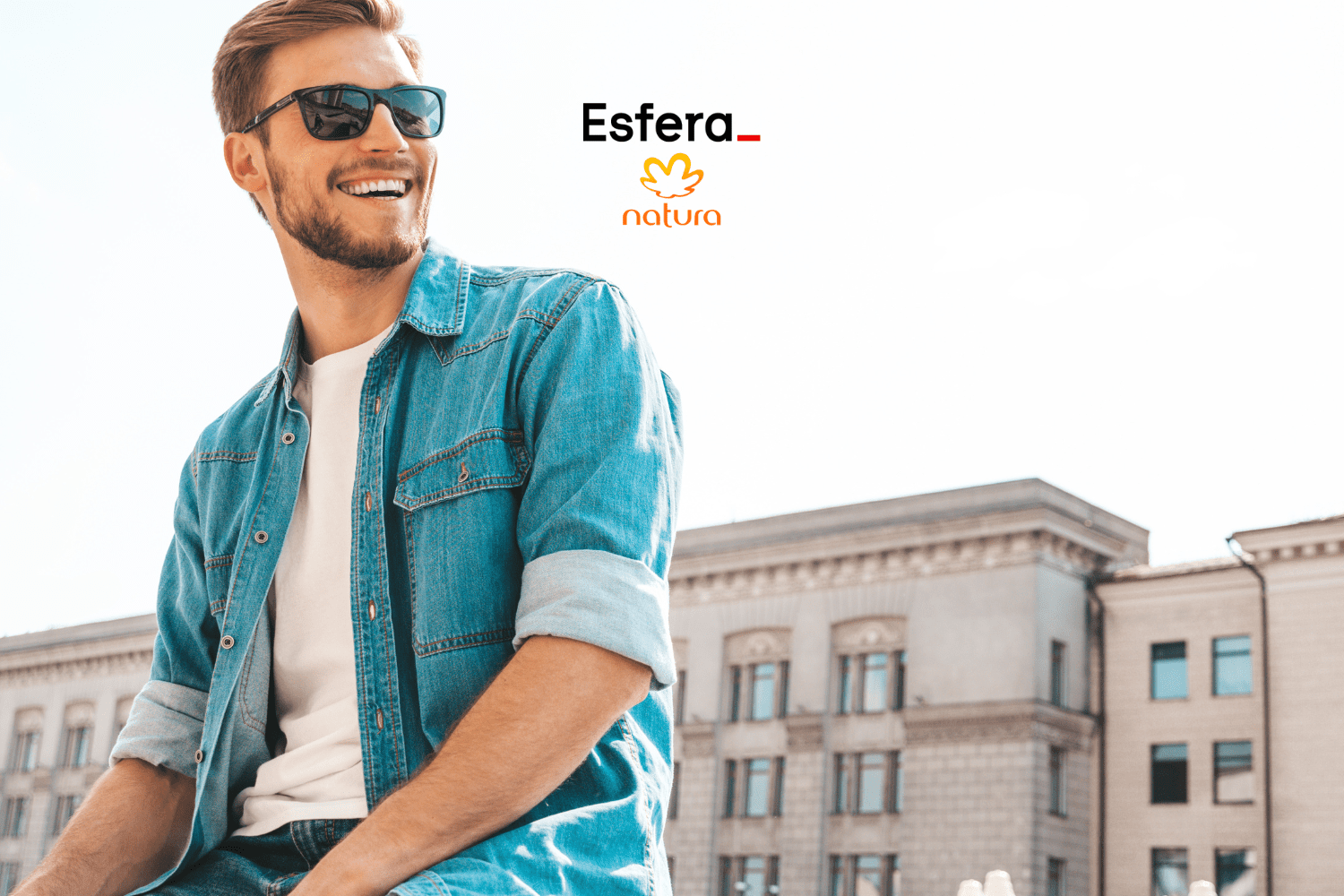 Homem branco sorrindo, usando óculos de sol, jaqueta jeans e camiseta branca, sentado ao ar livre em frente a um prédio histórico. Acima dele estão os logos da Esfera e da Natura, indicando uma colaboração entre as marcas.