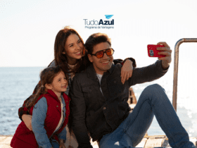 Descrição da imagem: Uma família com pai, mãe e filha pequena, sorrindo e tirando uma selfie à beira do mar, em um dia ensolarado. Acima deles, está a logo do programa de vantagens TudoAzul.